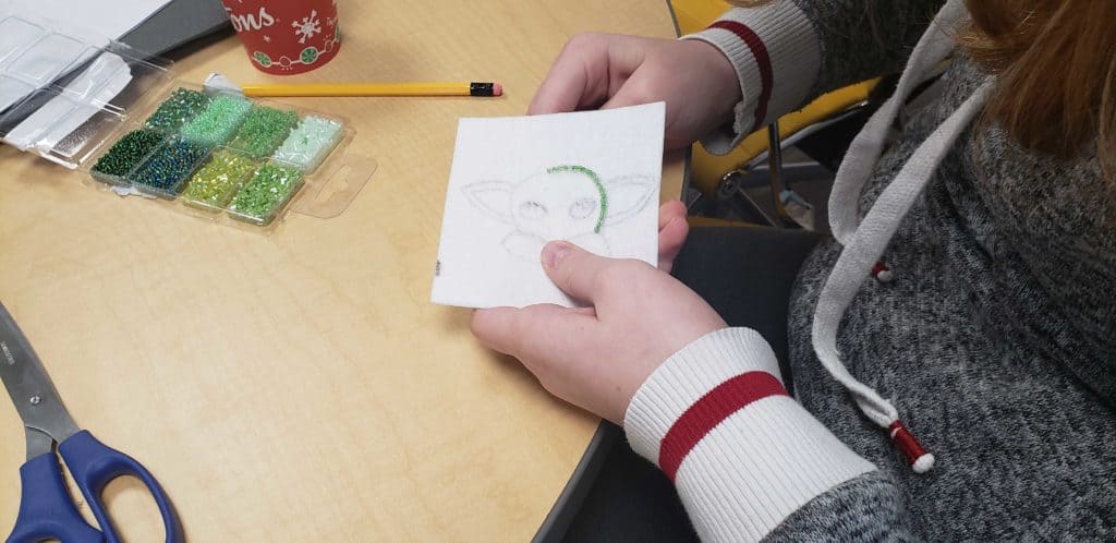 Someone is holding a patch of fabric that they are beading onto. There are scissors to the right bottom corner and beads placed above it.