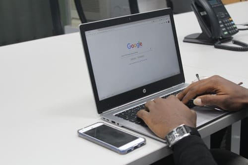 Hands on a laptop keyboard, there is an iPhone sitting beside the laptop.