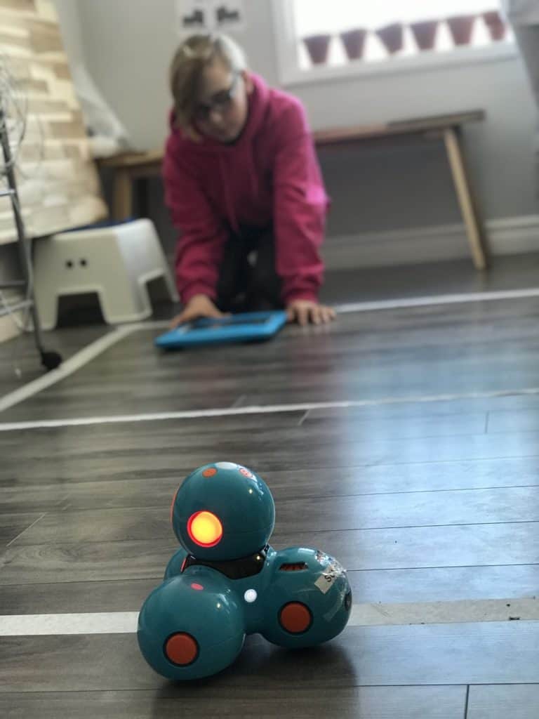 A Dash and Dot robot in main focus, with a student sitting on the floor with a tablet in the distance.