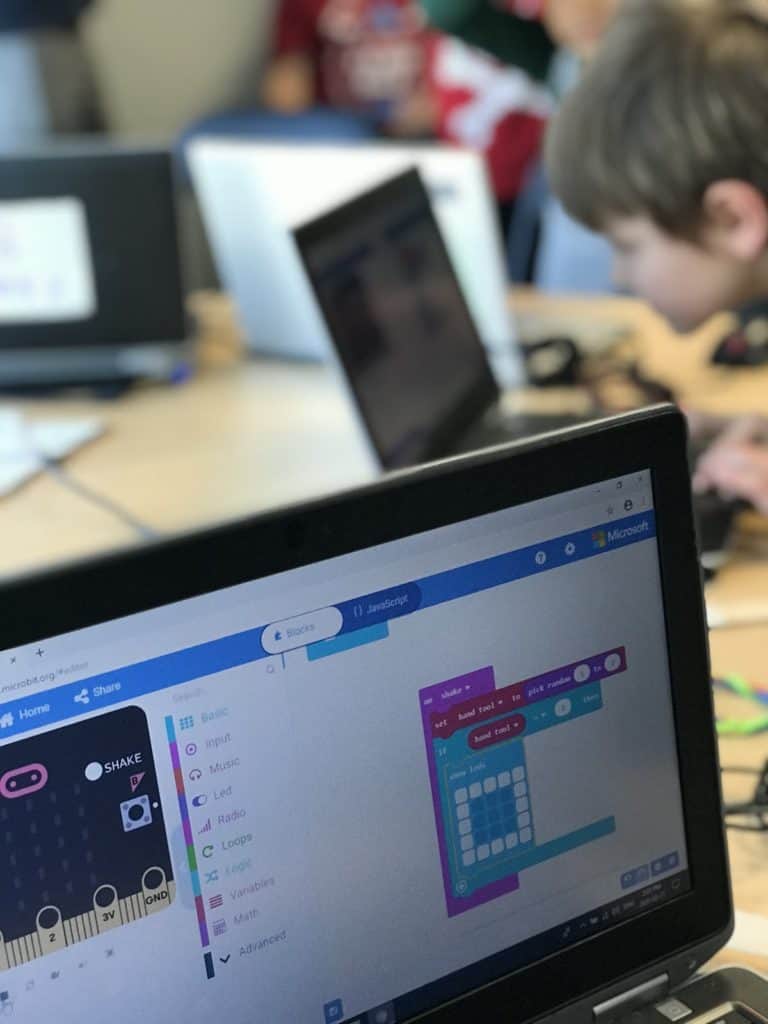 A computer screen open to a Scratch project in the main focus, as students work on their laptops in the background.