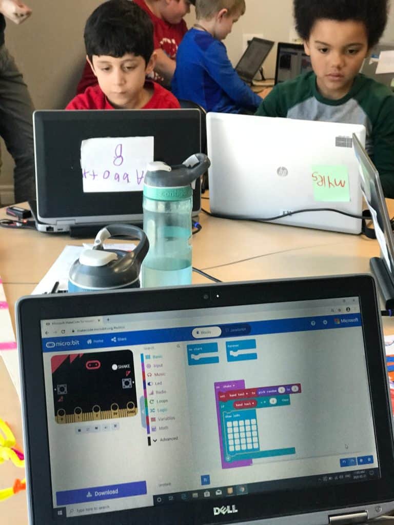 One computer screen facing the camera with Scratch open. Two students sitting at the same table, in the back, working on their computers.