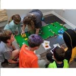 5 kids kneeling around a mat and building a lego obstacle courses for robots with an instructor kneeling beside