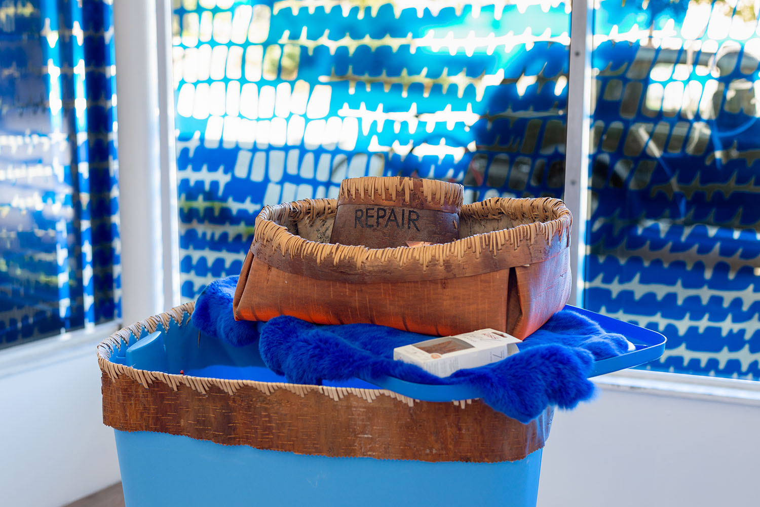 Two woven baskets, one small and one medium sized, sit on a big blue bin stitched along its rim with the same woven basket material. The small woven basket has the word "REPAIR" in black letters printed on the front of it.