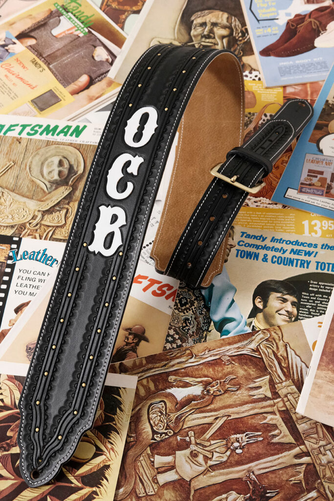 A custom black leather guitar strap embossed with white letters "OCB" lays atop a spread of vintage magazine pages.