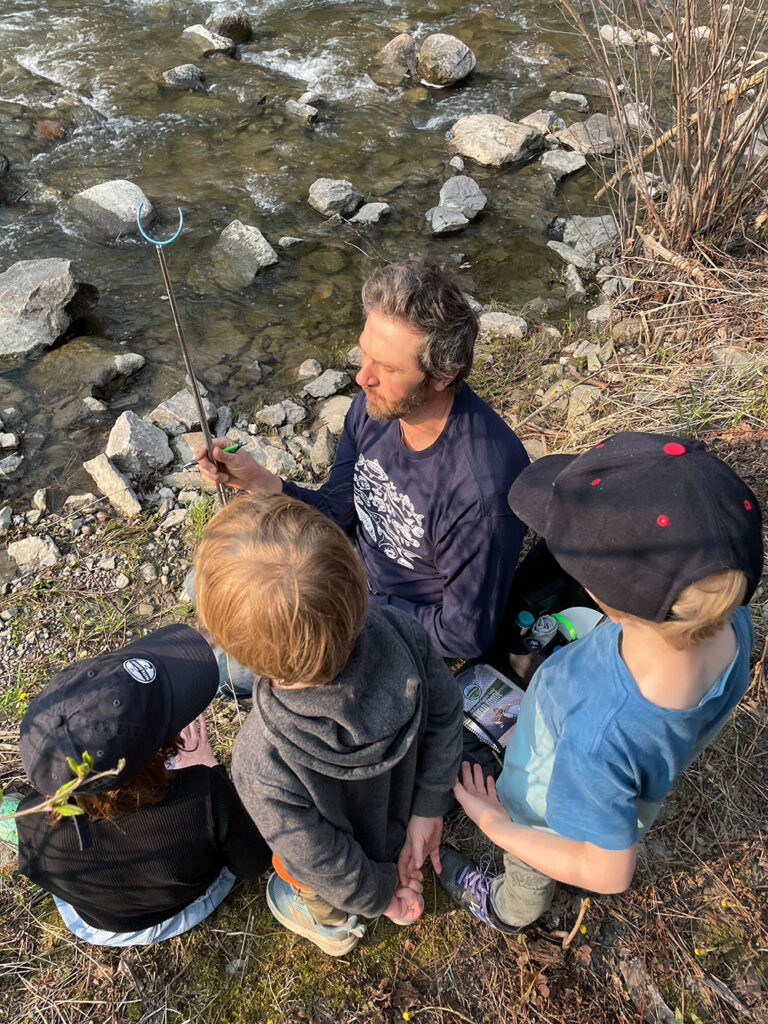 An instructor and three children sit by a river as they show them the grabber that comes in a water quality testing kit.
