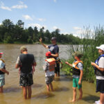 An instructor shows a group of children wearing life vests standing in a river how to use a water testing kit.