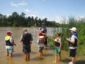 An instructor shows a group of children wearing life vests standing in a river how to use a water testing kit.