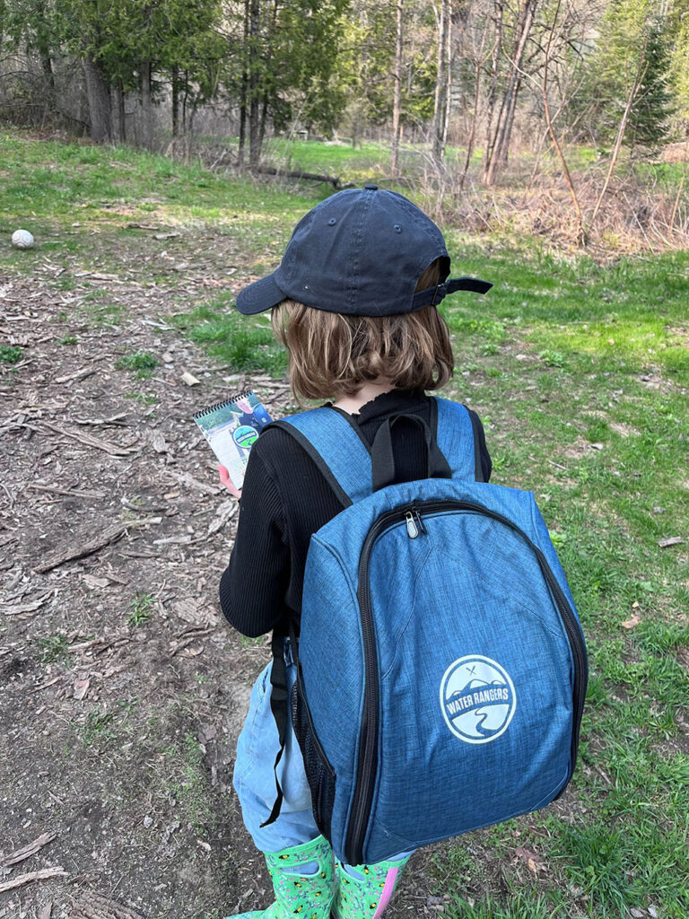 A child walks through a wooded area looking at a notebook and wearing a blue backpack with a logo and the words "Water Rangers" on the front of it.