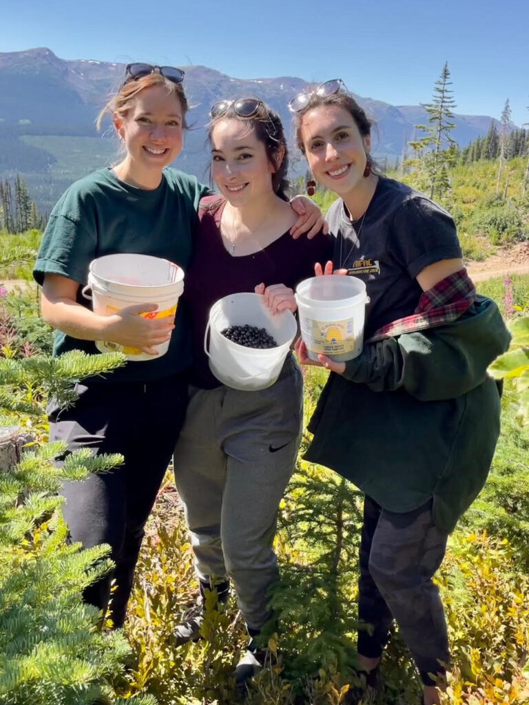 Three people stand together in a forested area, holding buckets filled with huckleberries.