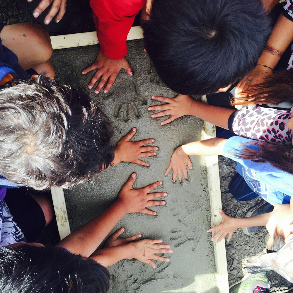 A birds-eye view of a group of children placing their handprints in a block of wet cement.